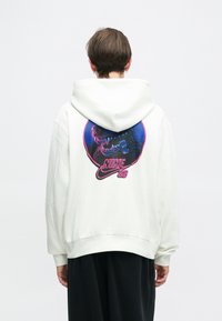 Sweatshirt branco com capuz, apresentando um gráfico colorido da face de um urso dentro de um design circular com o texto "NIKE SB". Tecido suave e texturizado.