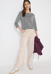 Femme portant un haut gris à manches longues et un pantalon beige à jambes larges, tenant une veste violette pliée dans une main, debout contre un mur blanc.