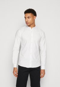 Camisa blanca de manga larga con cuello mao, que presenta un cierre frontal con botones y una textura suave, combinada con pantalones oscuros.