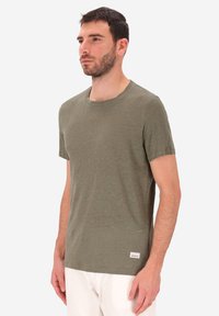 Uomo che indossa una maglietta verde oliva a maniche corte e pantaloni bianchi, in piedi con la mano sinistra rilassata al fianco, guardando leggermente a sinistra.