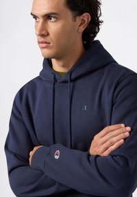 Felpa blu navy in tessuto morbido con tasca a marsupio e logo sul petto. Presenta un piccolo logo decorativo sulla manica.