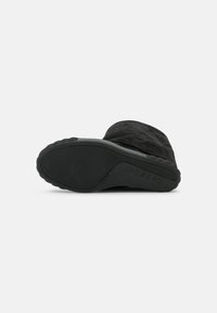 Bota negra de ante con suela de goma texturizada. La parte inferior presenta un diseño plano con un logo sutil y patrones de agarre para mayor tracción.