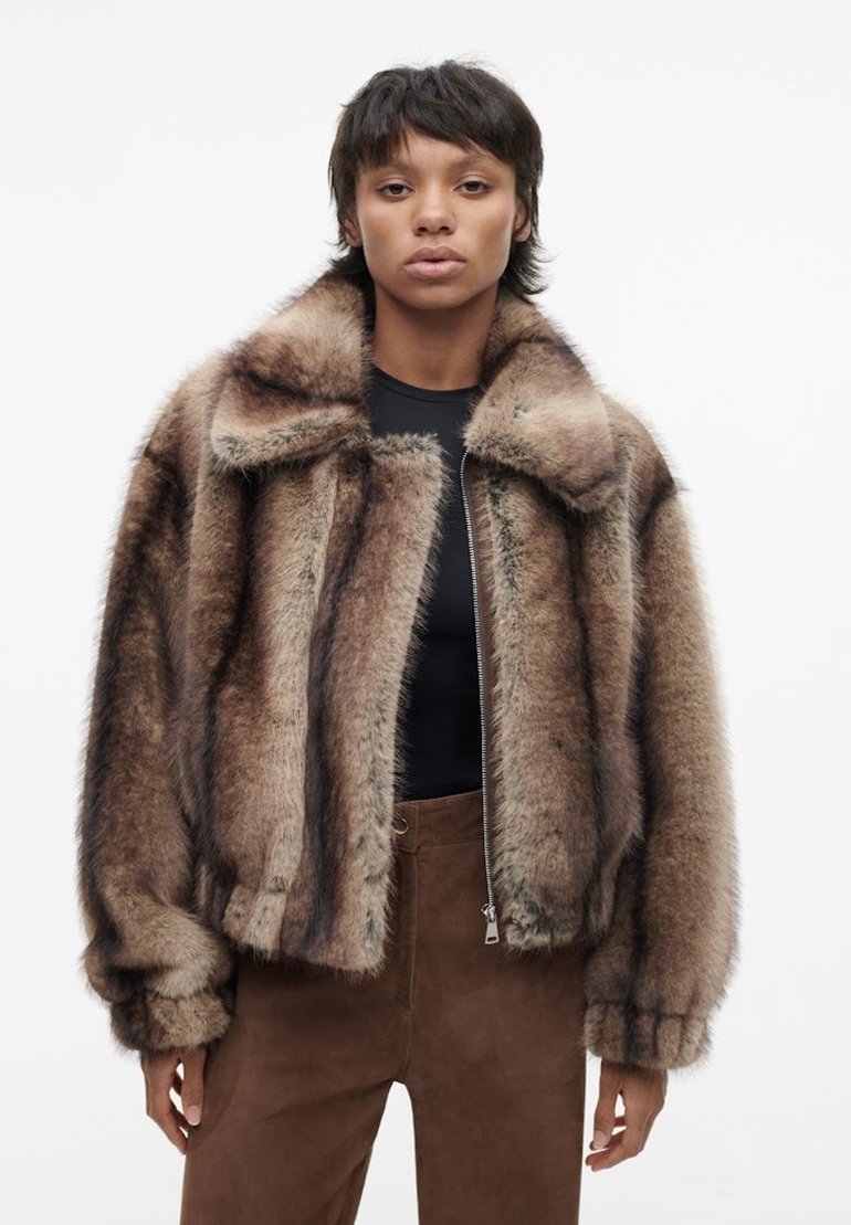 Vrouw in een bruin en zwart gestreepte nepbontjas over een zwarte top en een bruin broek, die naar voren kijkt tegen een effen witte achtergrond.