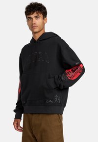 Schwarzes Hoodie mit Kängurutasche, ausgestattet mit einem großen Textlogo auf der Brust, einem roten Tiger-Graphic auf dem Ärmel und grauen Details am Saum.