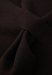 Gros plan d'un tissu brun foncé avec une surface texturée, montrant des plis intricats et un détail en forme de nœud.
