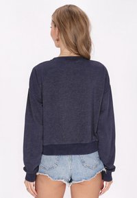 Felpa corta navy con maniche lunghe, vestibilità rilassata, polsini e orlo a costine, abbinata a pantaloncini di denim chiaro. Tessuto morbido e testurizzato.