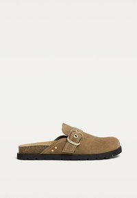 Brown suede slip-on clogs with an adjustable strap, flat black outsole, and textured footbed. Features a silver buckle accent.
