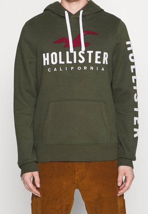 Felpa con cappuccio verde oliva con lacci bianchi, grande logo "Hollister" bianco e rosso, e tasca a marsupio. Testo bianco sul maniche.