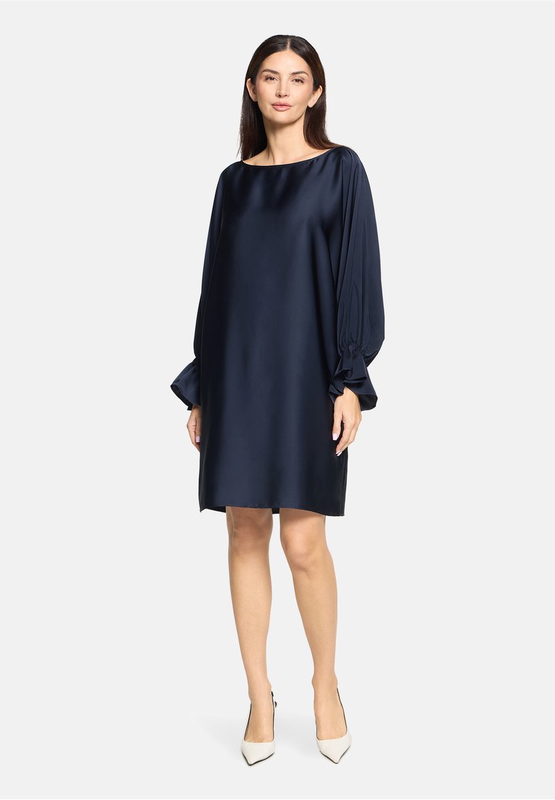 Robe en satin bleu marine avec une coupe loose, un large décolleté bateau et des manches plissées volumineuses, s'arrêtant juste au-dessus du genou. Les talons sont de couleur blanc cassé.