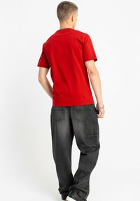 Czerwona bawełniana koszulka z krótkimi rękawami, zestawiona z czarnymi spodniami cargo z bocznymi kieszeniami i luźnym krojem. Widok od tyłu ukazujący prosty design.