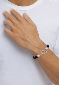 Svart flätad snoddarmband med silvriga detaljer och en rund låsning, prydd med en liten berlock. Bärs på en hand mot en vit skjorta.