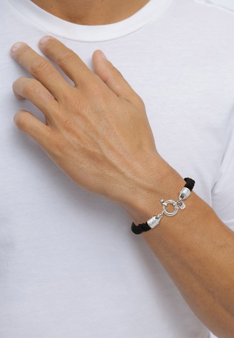 Svart flätad snoddarmband med silvriga detaljer och en rund låsning, prydd med en liten berlock. Bärs på en hand mot en vit skjorta.