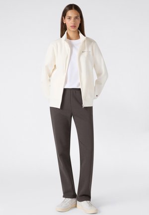 Mujer de pie con una chaqueta blanca con cremallera sobre una camisa blanca, pantalones gris oscuro y zapatillas blancas sobre un fondo claro y liso.