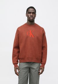 Vīnogu krāsas adīts sporta krekls ar ribveida apkakli un apakšējo malu, uz kura izšūts "CK" logotips sarkanā kontrastējošā krāsā. Krekls kombinēts ar pelēkām biksēm.