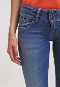 Detailní záběr osoby, která má na sobě modré džínové kalhoty se dvojitým knoflíkovým zapínáním a oranžové žebrované tričko, ruka ponechána volně podél těla.