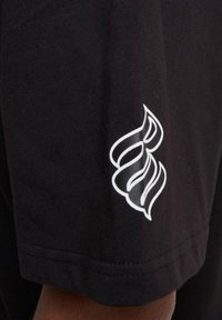 Juodas marškinėliai su baltu stilizuotu logotipu ant kairiojo rankovės, pagaminti iš minkštos medvilnės medžiagos su lygiu paviršiumi.