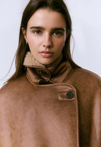 Mujer joven con cabello largo y marrón, vestida con un abrigo de cuero forrado de borreguito marrón con botones negros, frente a un fondo sencillo.