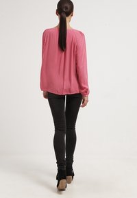 Růžová blůza s dlouhými rukávy a volným střihem, která má jemný lesk a na ramenou uchycené detaily, spárovaná s černými skinny džínami a botami na podpatku.