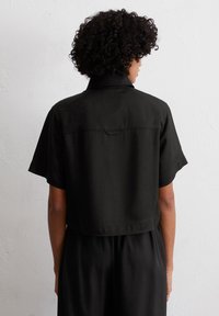 Zwart cropped shirt met korte mouwen, voorzien van een gladde textuur en een rug yoke detail. De stof lijkt licht en casual.