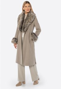 Světle šedý vlněný kabát se širokým chlupatým límcem a manžetami. Má dvouřadý design a boční rozparky. Nosen s béžovými kalhotami a botami.