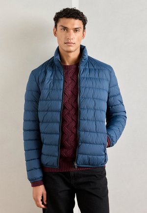 Blue padded jacket with a high collar, zippered front, and horizontal quilted design, worn over a burgundy cable knit sweater.