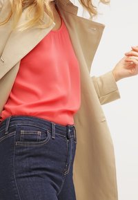 Top in satin corallo sotto un trench beige, abbinato a jeans di denim blu scuro. L'outfit presenta una vestibilità rilassata e dettagli di cucitura a vista.