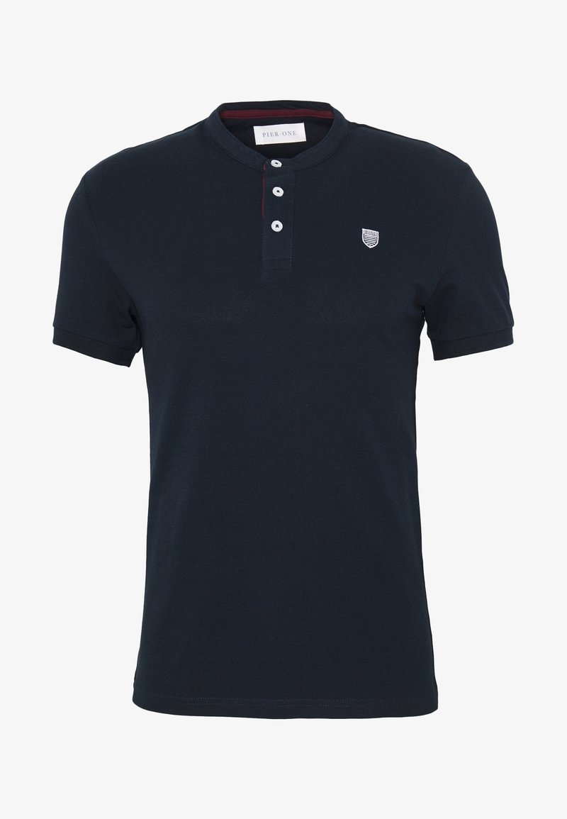 Polo bleu marine à manches courtes avec col rond, trois boutons blancs et un petit logo blanc en forme de bouclier sur la poitrine gauche.