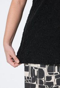 Černý top s texturovaným květinovým vzorem; látka vypadá jemně. Ruka modelky drží lem. Spodní část má abstraktní černobílý design.