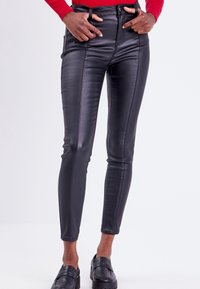 Pantalons skinny en simili cuir noir avec taille haute, poches avant et coutures distinctes. Texture lisse avec une finition brillante.
