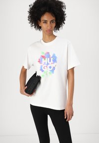 Tricou alb din bumbac cu un grafic abstract colorat și textul "HUGO", asociat cu accesorii negre și colanți negri. Textură netedă.