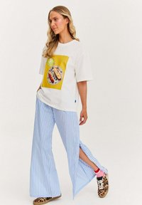 Bílý tričko s barevným potiskem jídla, doplněné modro-bílými pruhovanými širokými kalhotami s bočními rozparky a teniskami s tygřím vzorem.