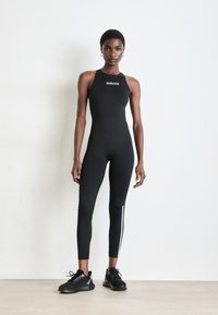 Zwarte mouwloze bodysuit met een hoge hals en een logo, voorzien van strakke pijpen en witte zijstrepen. In combinatie met zwarte sportschoenen.