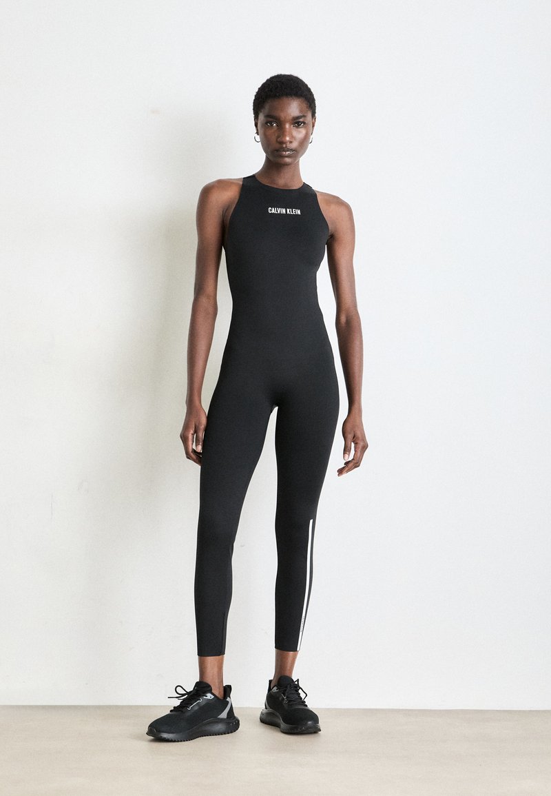 Zwarte mouwloze bodysuit met een hoge hals en een logo, voorzien van strakke pijpen en witte zijstrepen. In combinatie met zwarte sportschoenen.