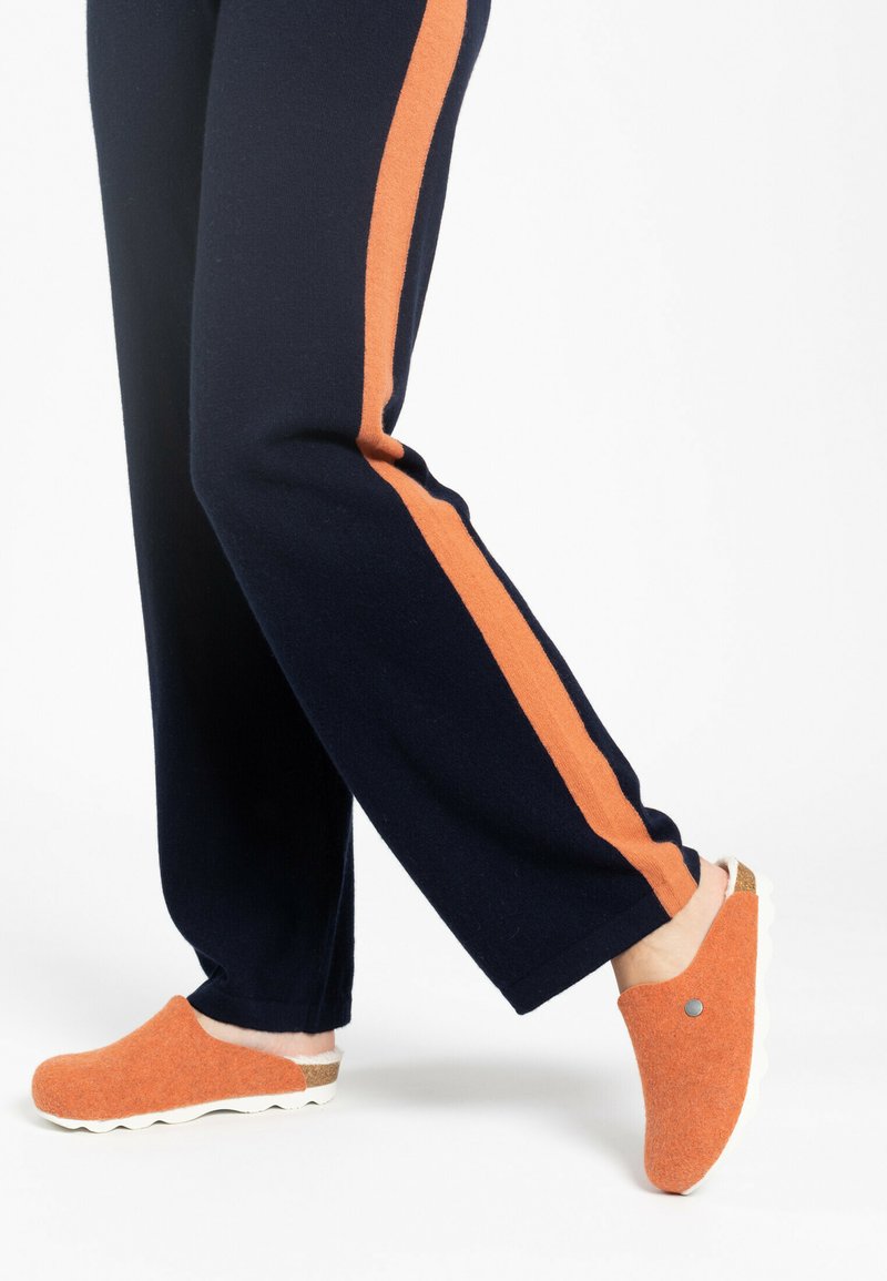 Zuecos de fieltro naranjas con suela blanca y acento de corcho. Se llevan con pantalones azul marino que presentan una franja lateral naranja.