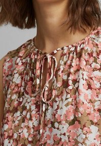 Femme portant un chemisier fleuri marron, rose et blanc avec un col rond et un détail noué à l'avant.