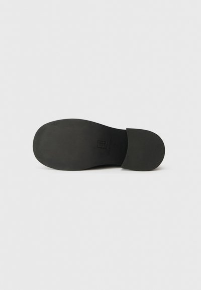 Μαύρη σόλα παπουτσιού slip-on με λείο ελαστικό υλικό, στρογγυλή μορφή και διακριτές λεπτομέρειες επωνυμίας. Ο σχεδιασμός είναι επίπεδος χωρίς ανύψωση τακουνιού.