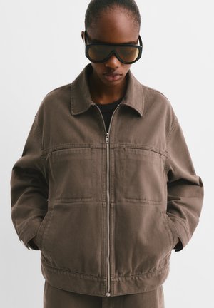 Brown canvas jacket with a zip front, large pockets, and a collar. Model wears oversized black sunglasses. Smooth texture with stitching details.