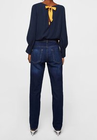 Blouse bleu marine à manches bouffantes, détail noué dans le dos ; associée à un jean droit bleu foncé et des chaussures blanches à talons hauts.