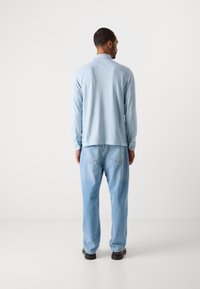 Mann som står med ryggen til iført en lys blå langermet skjorte, lyseblå jeans og svarte sko mot en ensfarget hvit bakgrunn.