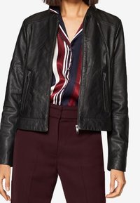 Mustaa nahkatakkia, jossa on lyhennetty muotoilu, vetoketjukiinnitys edessä ja kaksi sivutaskua. Sileä pinta, minimalistinen varuste ja ei näkyviä koristeluja.