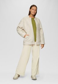 Beige Jacke aus Wollmischung mit texturierter Oberfläche, oversized Schnitt, zwei vordere Taschen und Druckknöpfen, kombiniert mit einem grünen Oberteil und cremefarbenen Hosen.