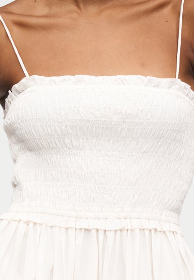 Femme portant une robe blanche froncée, bustier avec fines bretelles spaghetti et un ourlet à volants sur la poitrine.