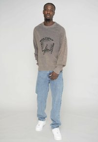 Übergroßer beiger Pullover mit gerippter Textur und grafischem schwarzen Aufdruck, kombiniert mit hellblauen baggy Jeans und weißen Turnschuhen.