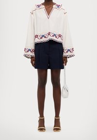 Persona vestida con blusa blanca bordada con motivos florales, pantalones cortos plisados en azul marino, sandalias beige con tiras al tobillo y sosteniendo un pequeño bolso tejida blanco.