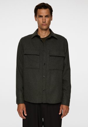 Veste chemise en laine mélangée vert foncé avec un col pointu, deux poches poitrine, et des boutons noirs. Forme structurée, texture lisse, manches longues.