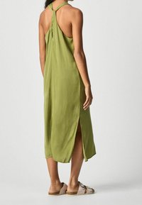 Žena oblečená v bezrukávových midi šatech olivově zelené barvy se spodním rozparkem a béžových sandálech s klínkem, stojící proti jednoduchému šedému pozadí.