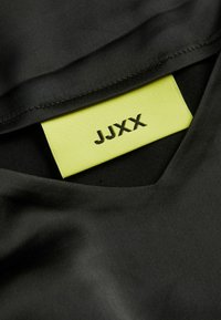 Tecido preto com uma textura suave, apresentando um rótulo verde que diz "JJXX" em texto preto, posicionado próximo ao decote em V.