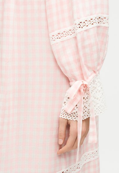 Χέρι ακουμπισμένο σε ροζ και λευκό γκίνχαμ ύφασμα με δαντελένιο τελείωμα και δεμένο φιόγκο στο μανίκι ενός μακριού ενδύματος.