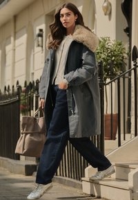 Manteau gris avec un col en fourrure, associé à un pull léger et un jean foncé. Tenant un sac beige, portant des baskets argentées. Décor urbain.