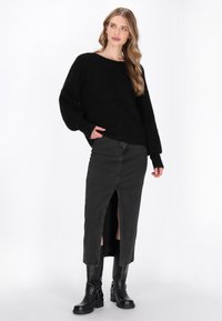 Schwarzer, locker sitzender Pullover mit langen Ärmeln, kombiniert mit einem schwarzen Jeansrock mit Schlitz vorne. Schwarze kniehohe Stiefel mit Riemen.